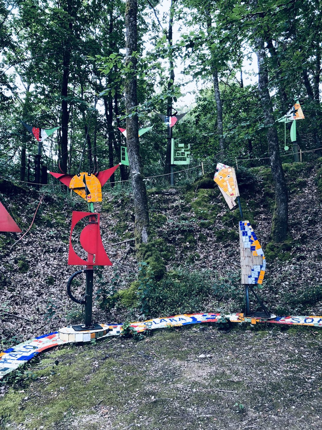 Un sentier boisé jalonné de totems mosaïqués et de visages tutélaires, chacun dédié à un nom gravé au sol.